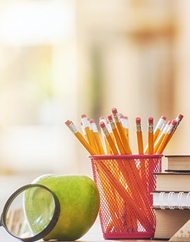 Pencils, an apple and a magnifying glass sit on a desk next to stacked books